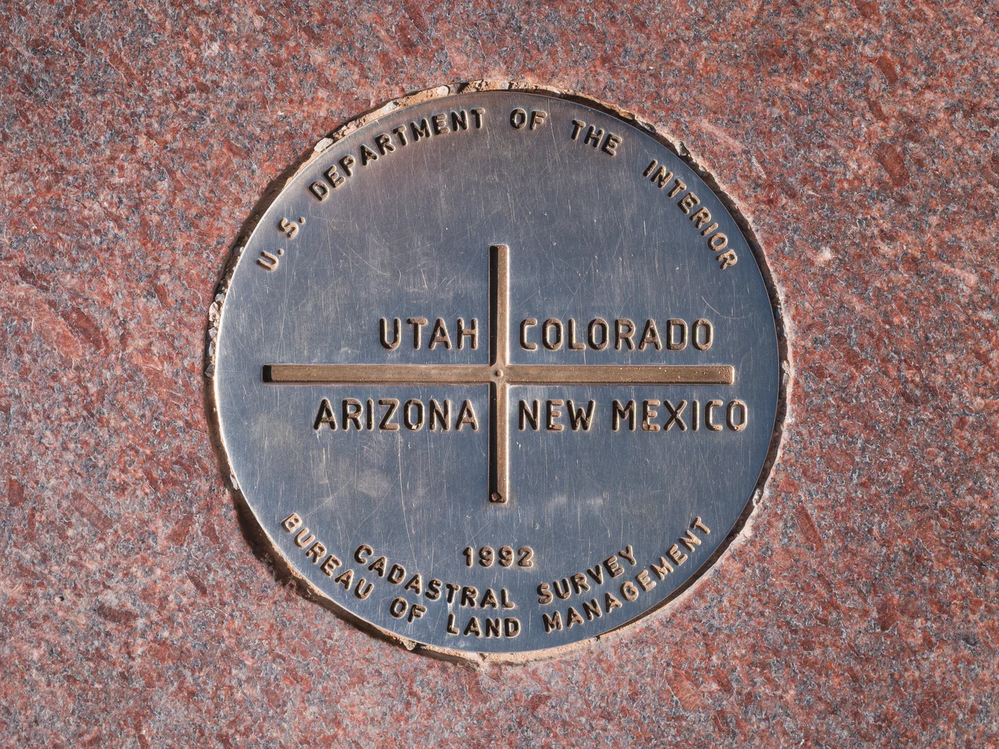 Le disque géodésique au centre du monument : une plaque de bronze encastrée à fleur du granit rouge poli, une croix en relief le divisant en quadrants portant les mots UTAH, COLORADO, ARIZONA et NEW MEXICO, et à son pied 1992 CADASTRAL SURVEY, BUREAU OF LAND MANAGEMENT