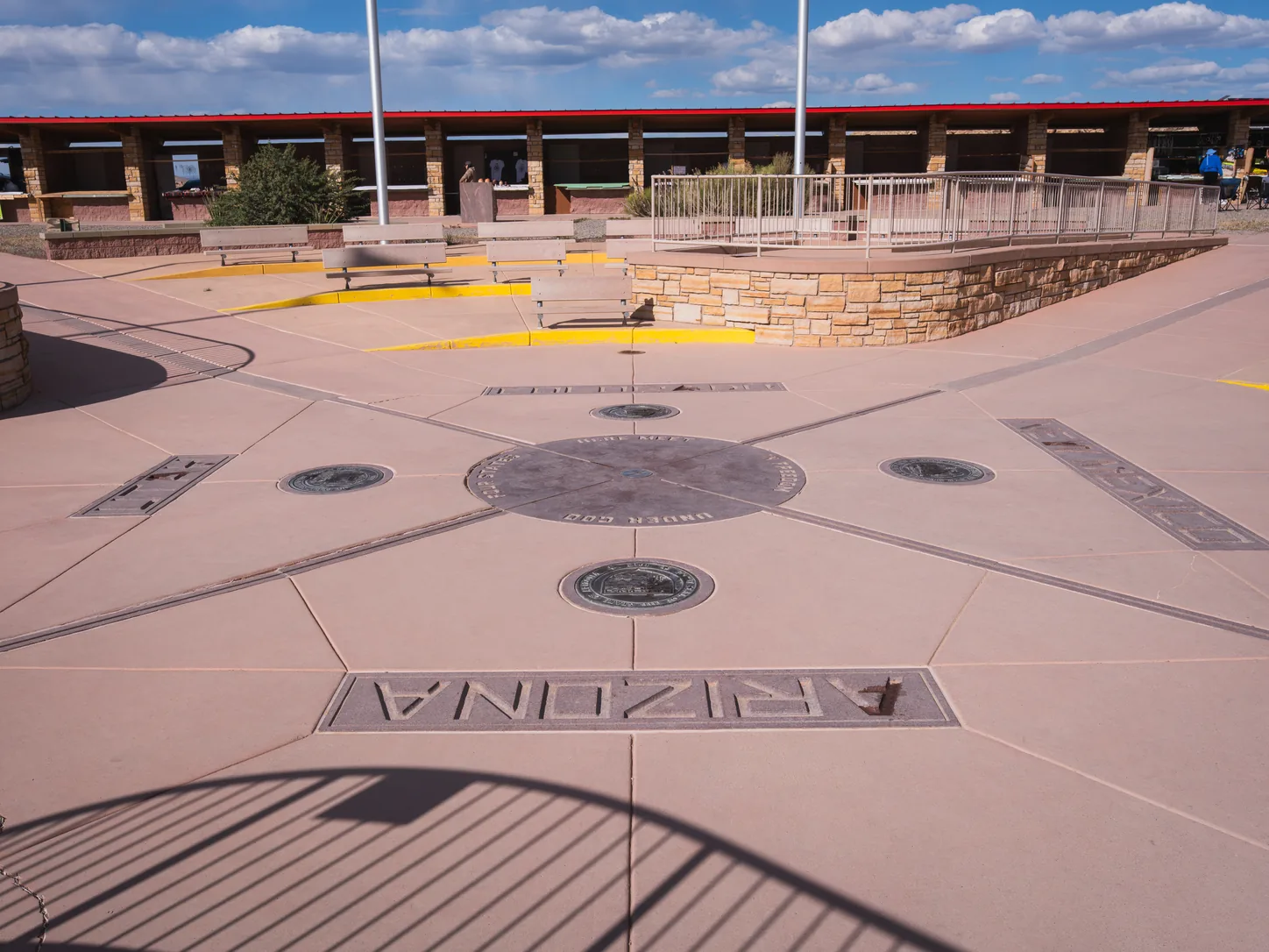 L’esplanade dallée et plate vue au ras du sol depuis le quadrant de l’Arizona, ARIZONA gravé dans le dallage au premier plan, les sceaux des États en bronze encastrés dans chaque quadrant, et les longues galeries d’artisanat ouvertes qui longent le côté opposé