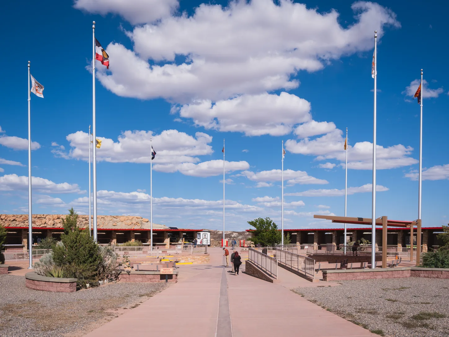 L’esplanade vue depuis l’allée sud, une rangée de mâts de drapeaux de chaque côté et les deux longues galeries ouvertes qui abritent les étals d’artisanat navajo et ute, avec le désert plat qui s’étend derrière